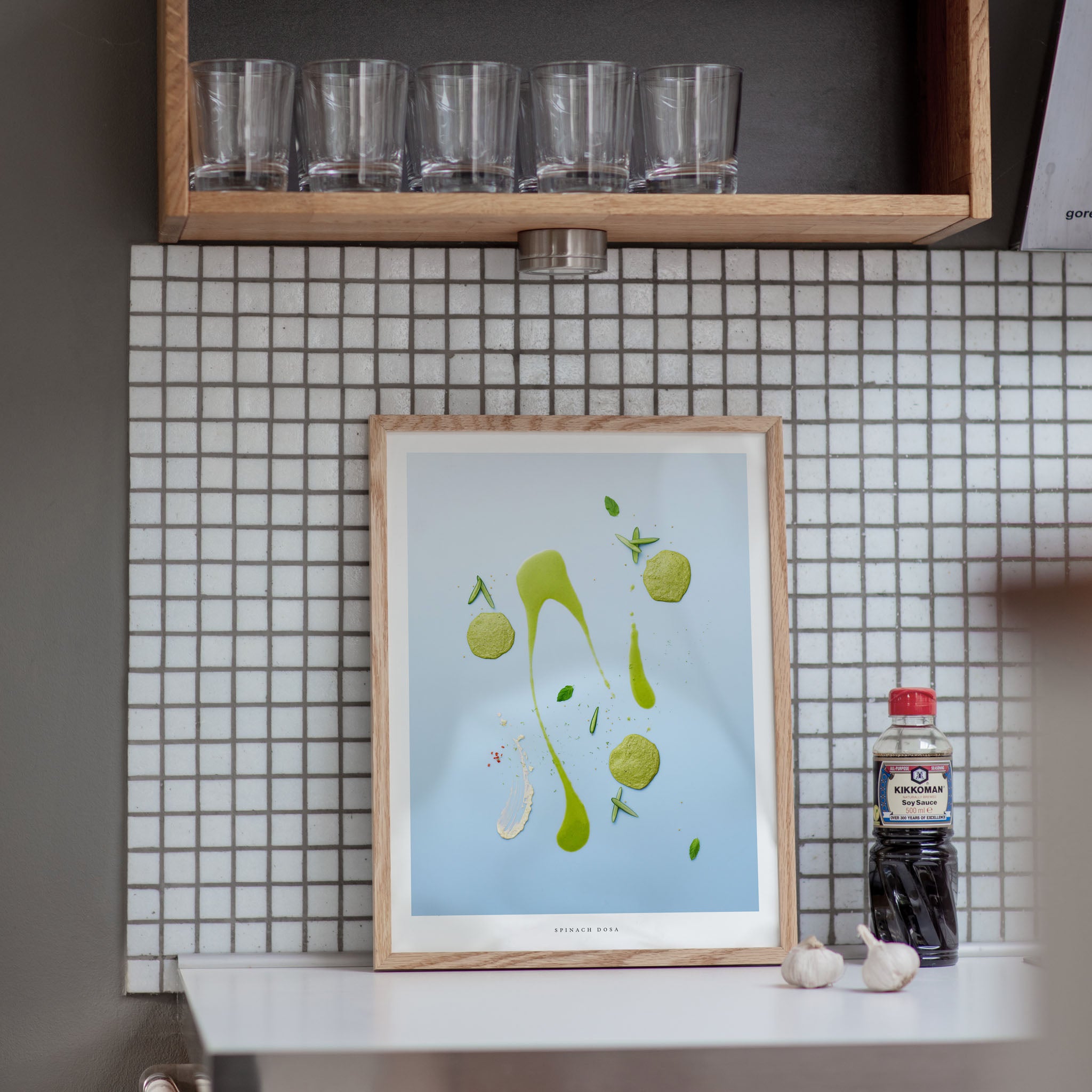 Framed artwork on a kitchen counter with tiled wall and shelf in the background