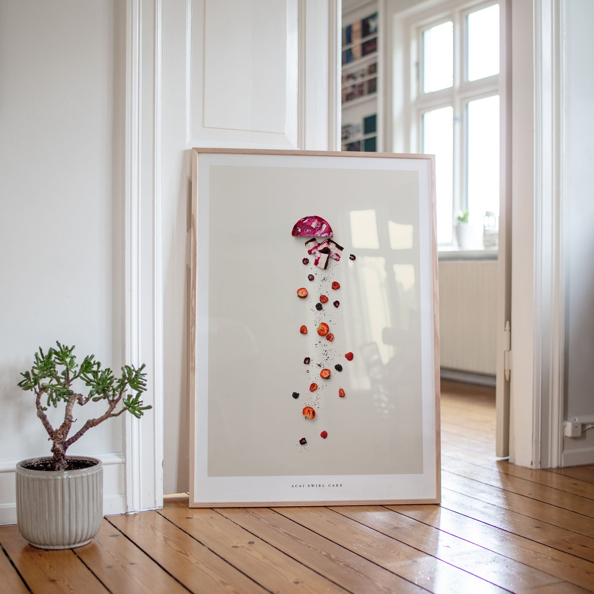 Framed artwork of a cake on a white wall in a room with wooden flooring and a plant.